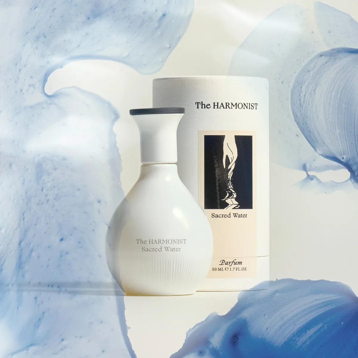 A white bottle of The Harmonist's "Sacred Water" perfume, paired with its packaging, set against a soft backdrop of flowing blue watercolor textures.