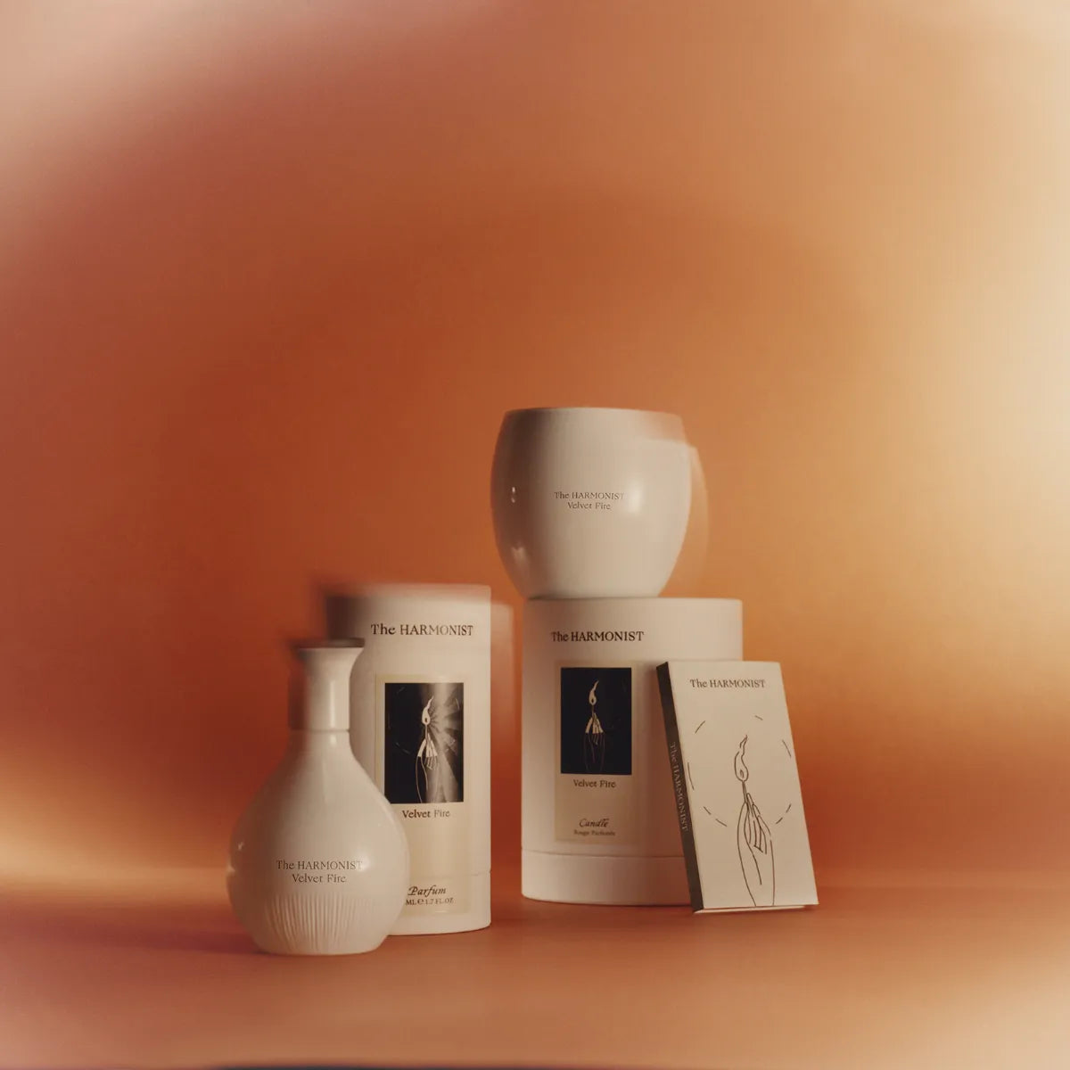 The Harmonist Velvet Fire collection featuring a white perfume bottle, candle, and packaging, set against a warm orange background.