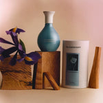 A perfume bottle and packaging of The Harmonist's "Yin Transformation" scent, styled with wood blocks and a purple iris flower against a soft, warm background.